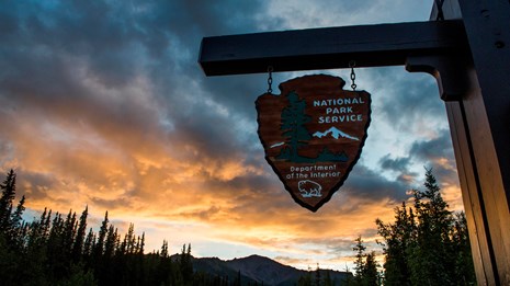 a sign with the words national park service hangs in front of a sky tinged yellow by setting sun