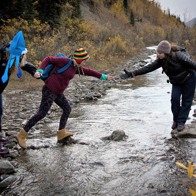 a young visitor crosses a stream with the helping hands of fellow hikers