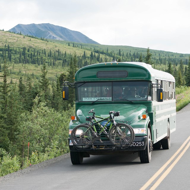 A green bus with a bike attached to the front drives down a paved road through a hilly landscape.