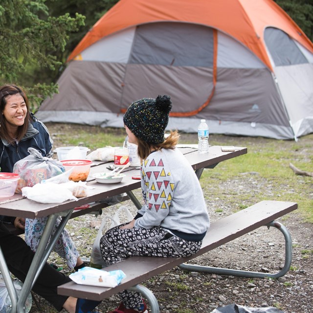 Three women sit at a picnic table, laughing and eating food. Behind them is a large orange tent.