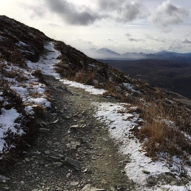 A dirt trail, with a fresh dusting of snow on the edges of the trail, leads up a mountainside.