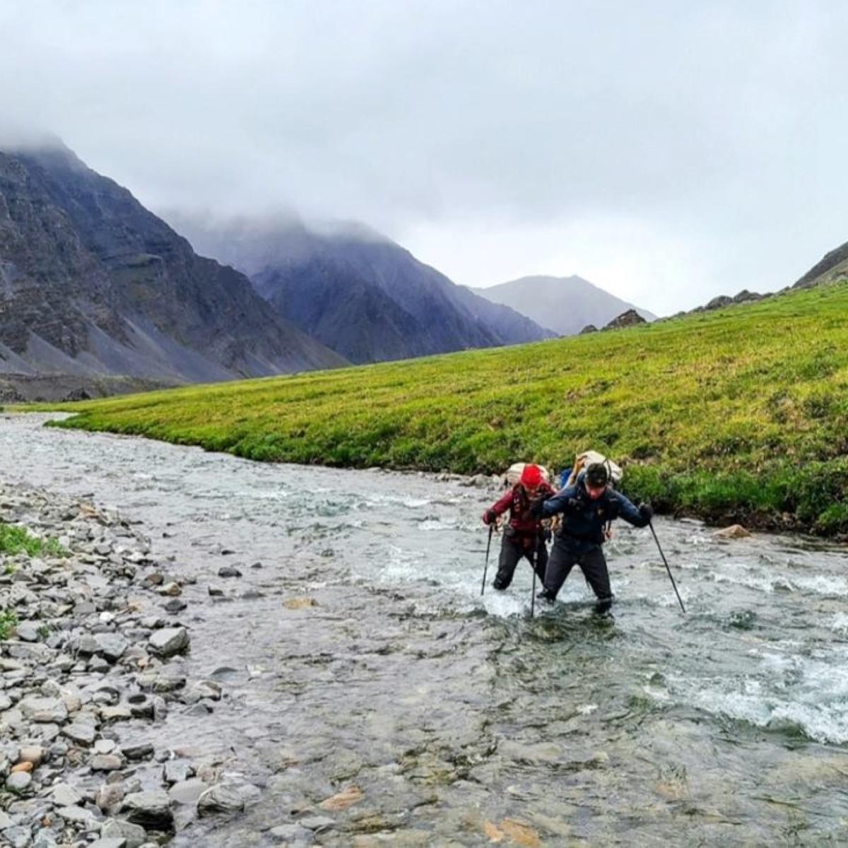 Two people wearing backpacks ford a river at the base of large, rocky mountains.