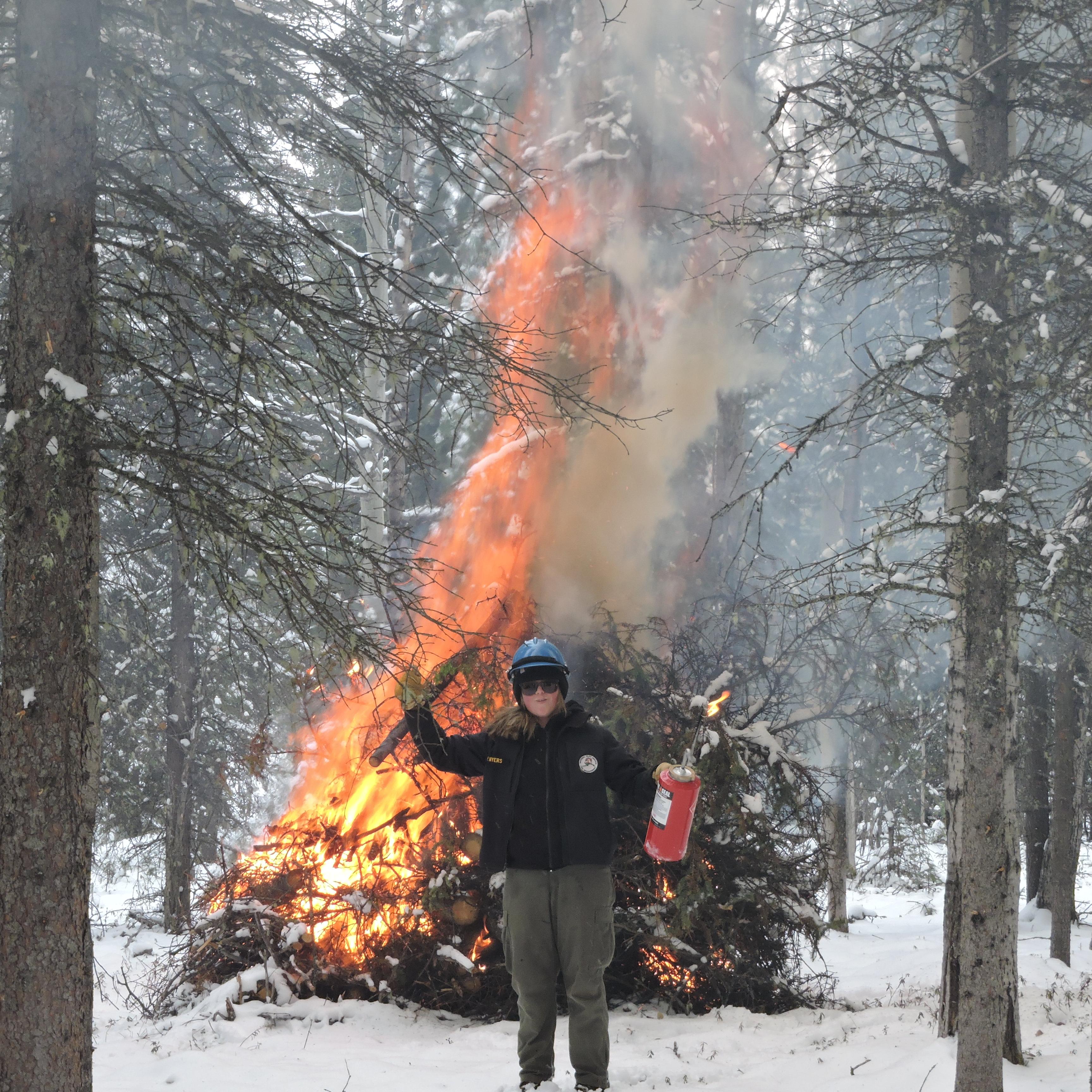 A woman in grey pants and a black shirt and jacket stands in front of a burning pile of brush.