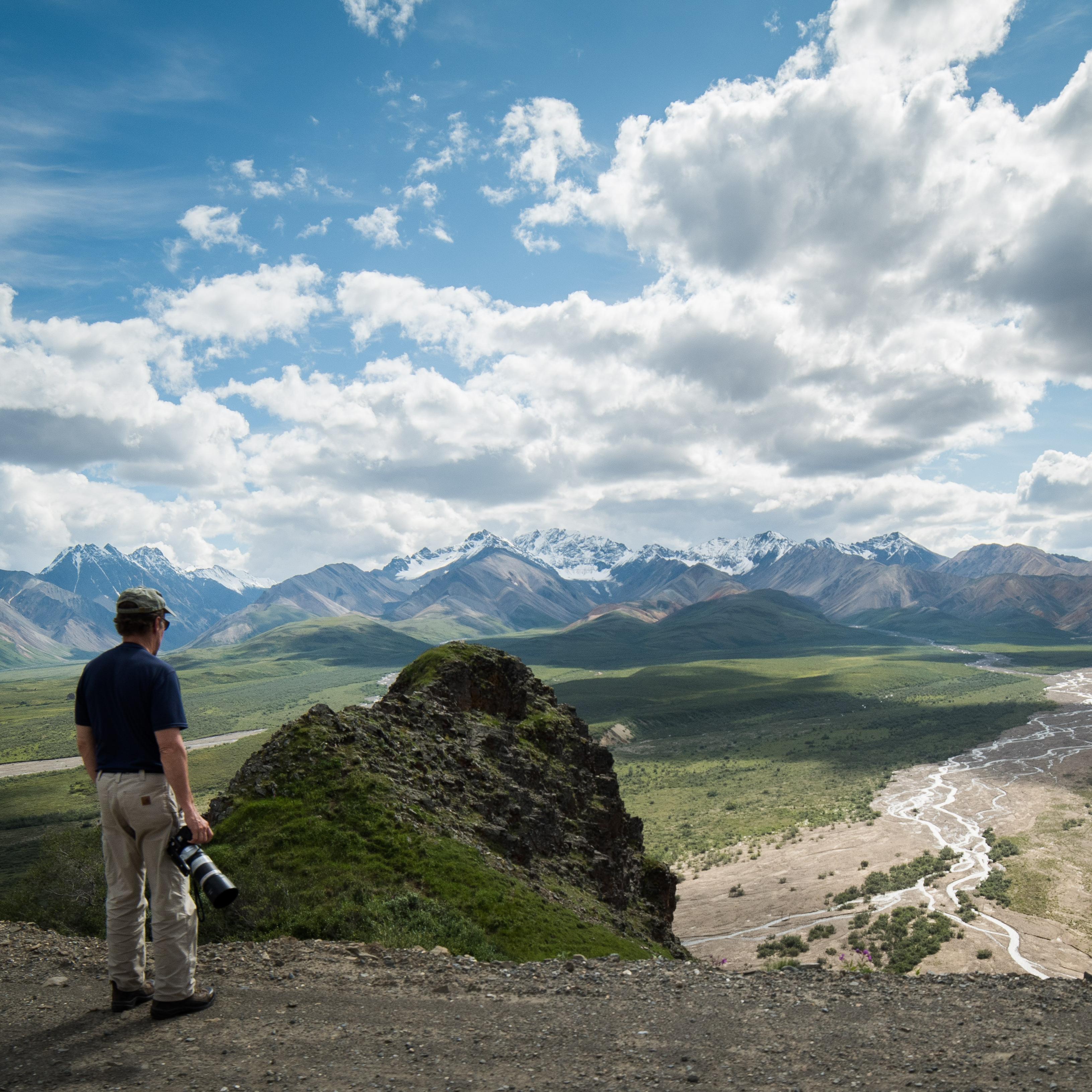 a man standing at a scenic overlook above a wide green plain leading up to distant mountains