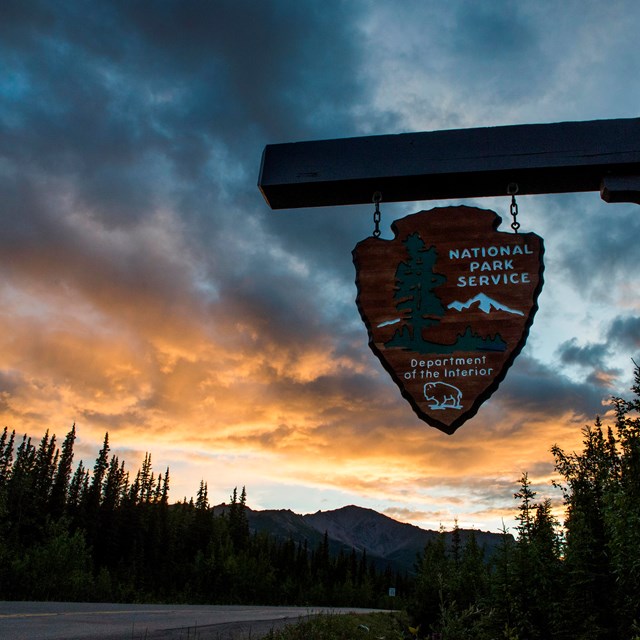 a sign with the words national park service hangs in front of a sky tinged yellow by setting sun