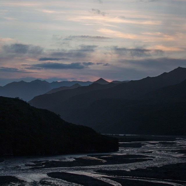 dark mountains and a shallow, braided river under a partly cloudy sky