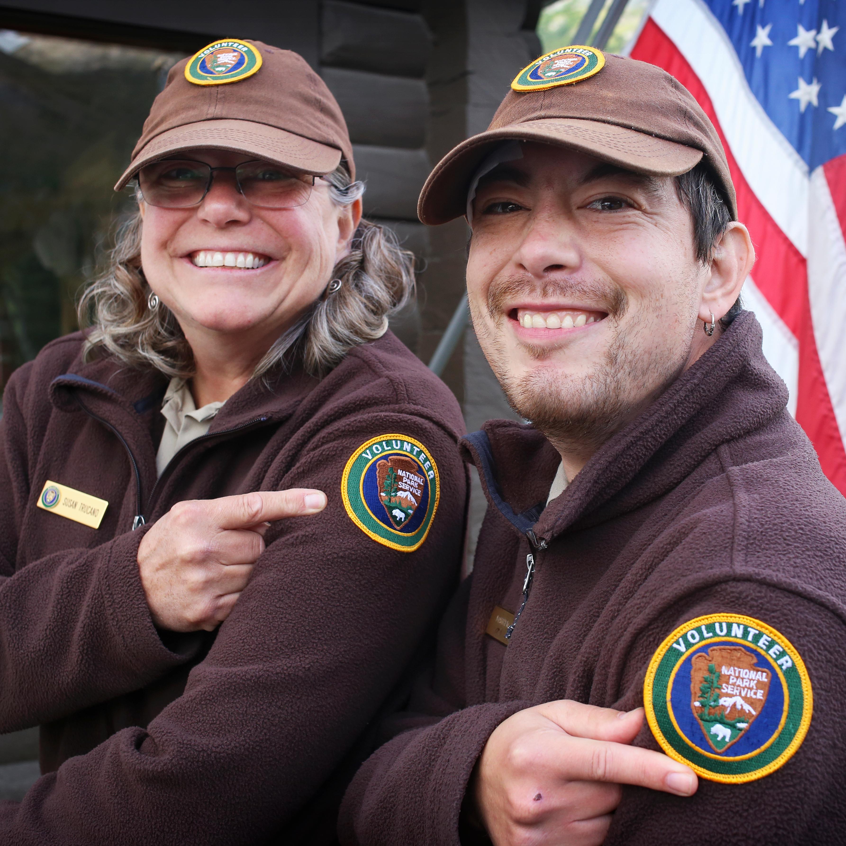 A smiling woman and man in volunteer uniforms point to the volunteer patch on their sleeves.