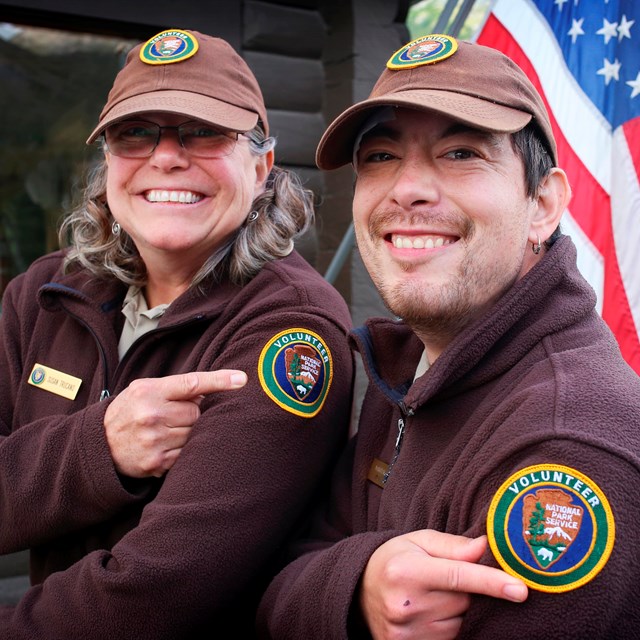 A smiling woman and man in volunteer uniforms point to the volunteer patch on their sleeves.
