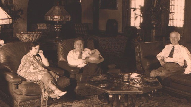 A woman sits with two men on leather couches in a grand living room. 
