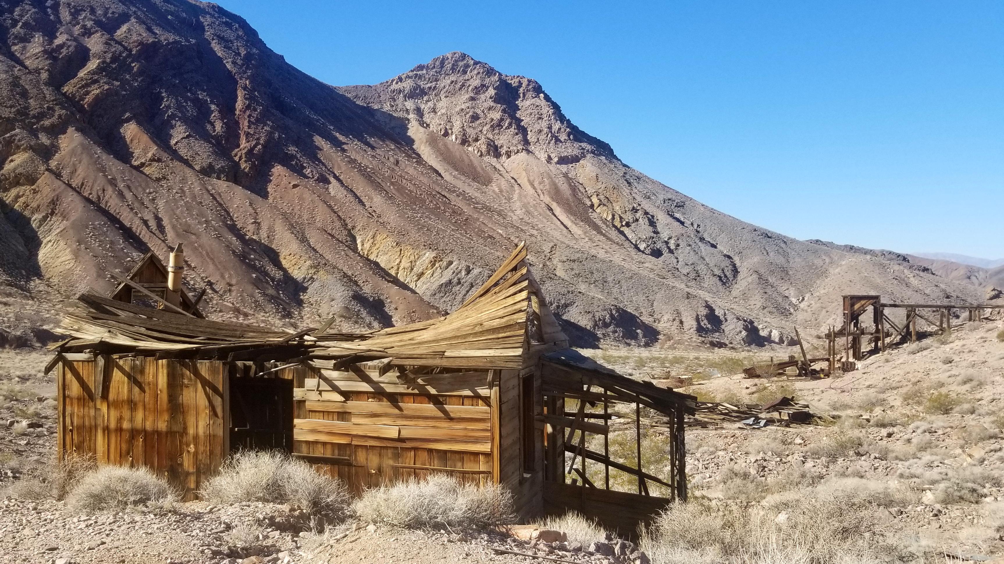 The remains of a weathered wooden building with a sagging roof stands before the remains.