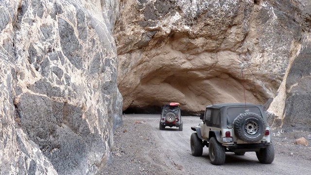 Two jeeps drive away from the camera through the marbled narrow rock walls of Titus Canyon.