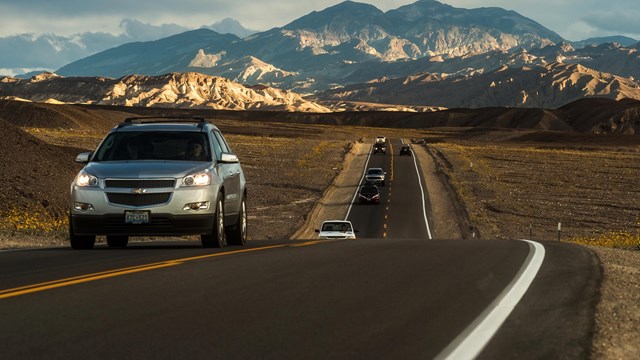 cars drive on a straight stretch of road in a desert setting with mountains in the distance