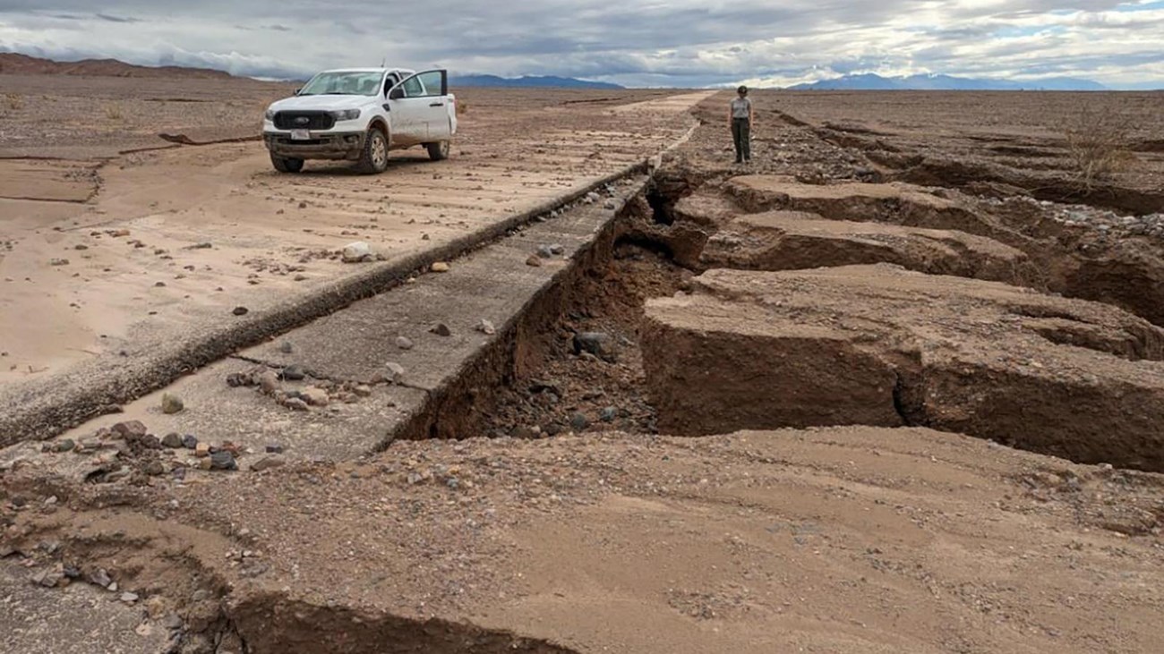 Ranger stands on mud cracks that extend deep into the earth next to a mud covered paved road.