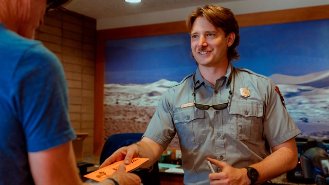 A smiling park ranger in uniform hands a paper pass or ticket to a visitor standing at a counter.