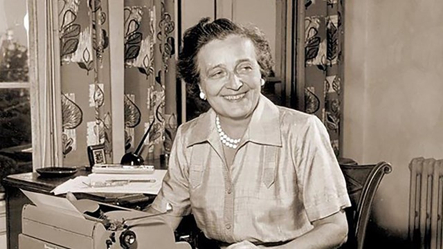 Sepia toned photo of woman in dress smiling seated at desk with typewriter.