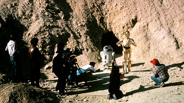 Two robots are surrounded by a few people filming them in a desert landscape. 