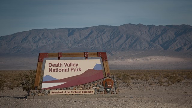 Death Valley National Park sign with distant, highly-eroded, brown desert mountains.