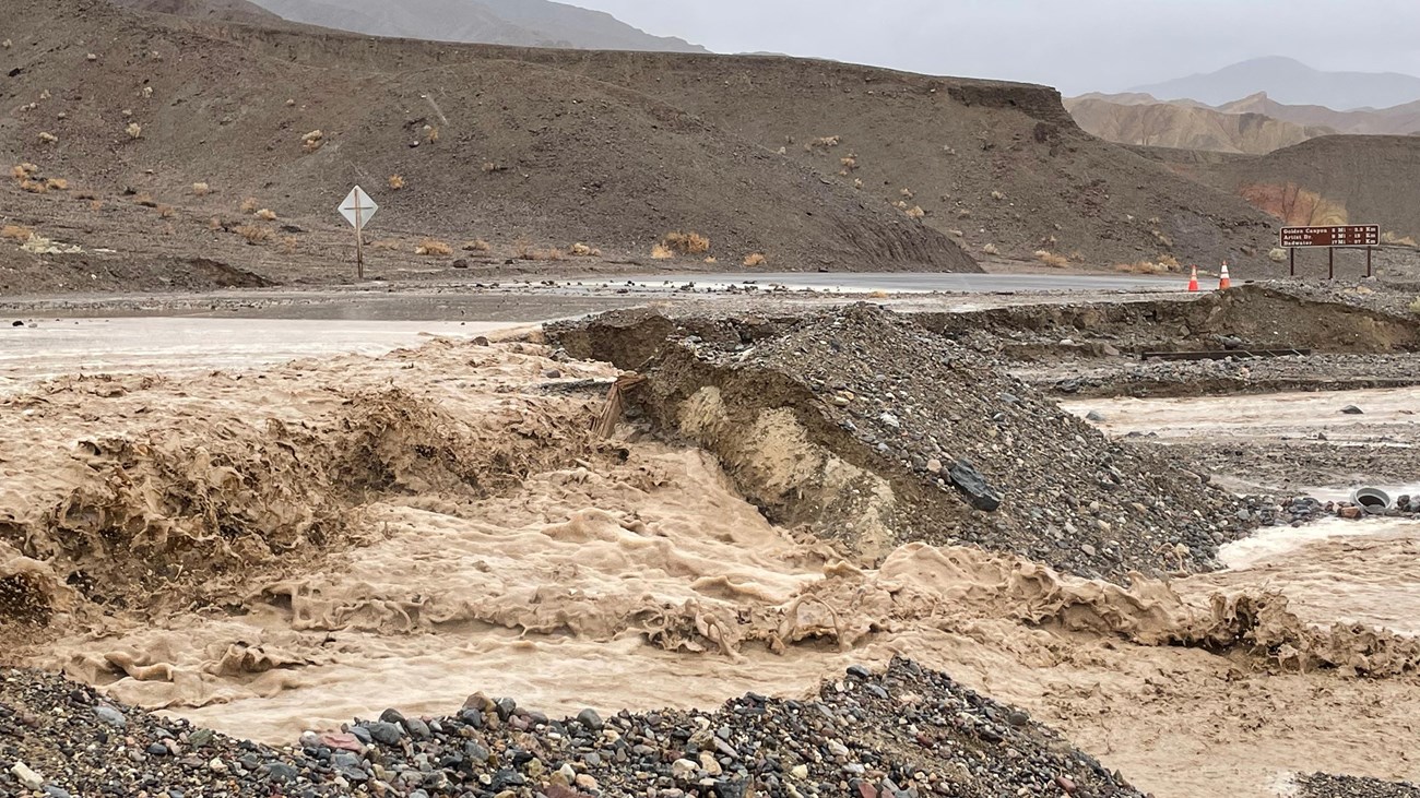 Muddy floodwater rushes over a paved road in a rocky desert and undercuts the downhill side.