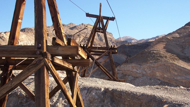 Two wooden towers connected near the top by metal cables in a hilly desert landscape.