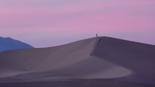 Warm, pink wispy clouds above a distant sand dune with a person on top and a blue mountain beyond. .