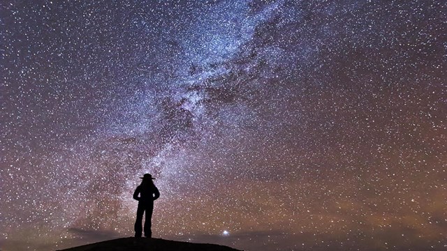A ranger silhouette stands in front of a milky way, starry backdrop. 