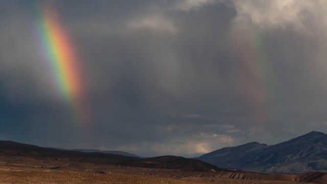 A double rainbow in a clouded sky, above a stark desert landscape. 