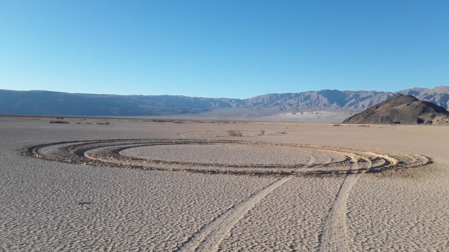 Straight and circular tire tracks on a brown desert playa with mountains in the distance.