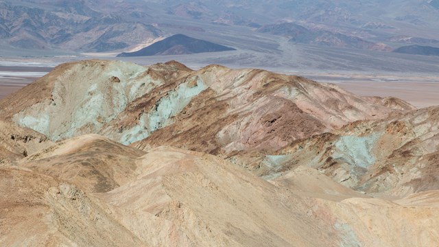 A mutli-hued, highly eroded hill with a horizon of desert mountains. 