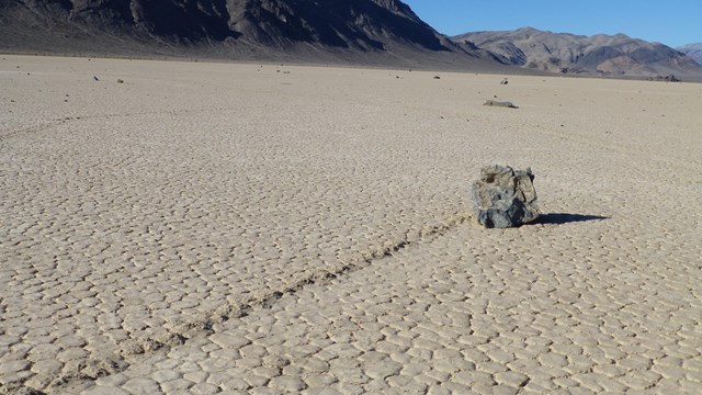 A track on a flat playa leads to a singular rock under a blue sky. 