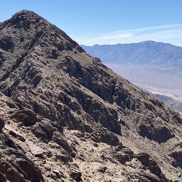 A rocky conical peak with desert mountains and valley in the distance.