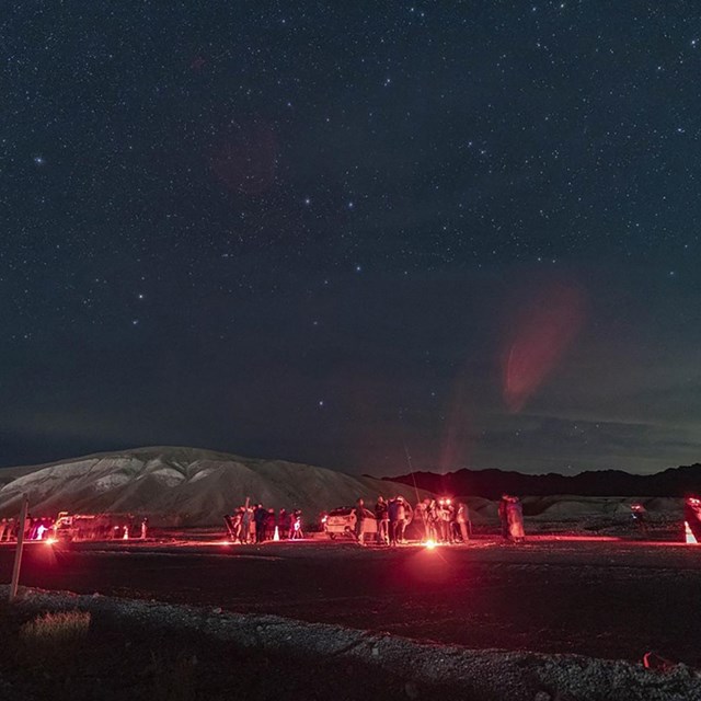 Various individuals use red light at night among many stars and a hilly landscape.
