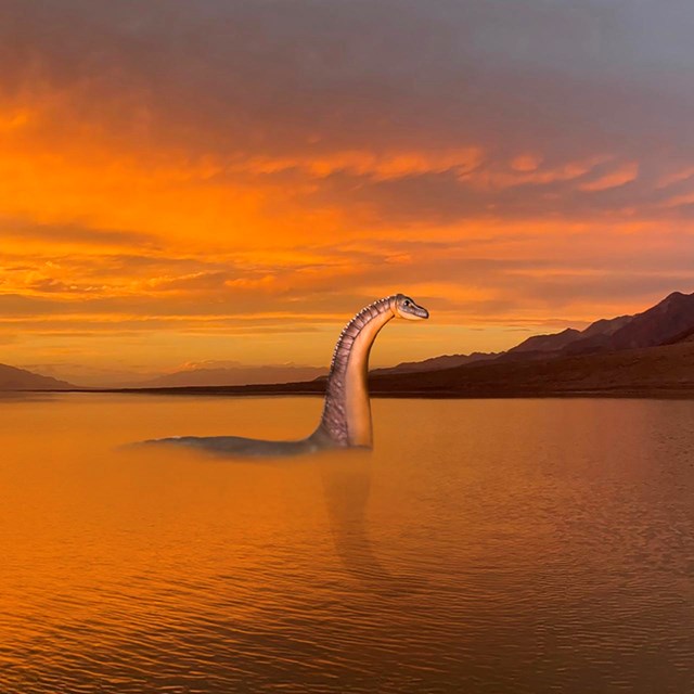 A fake reptilian creature standing mostly submerged in water against a cloudy orange sky.