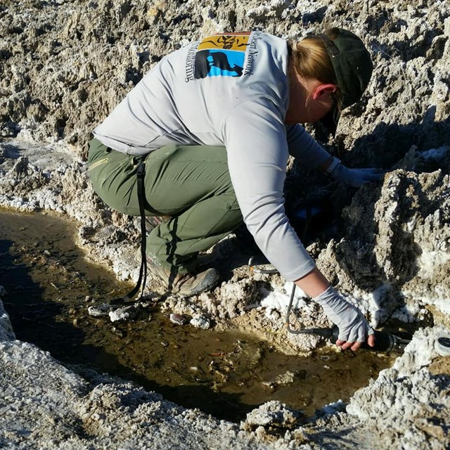 Woman kneeling by small spring in barren desert environment, holding a data sensor in the water