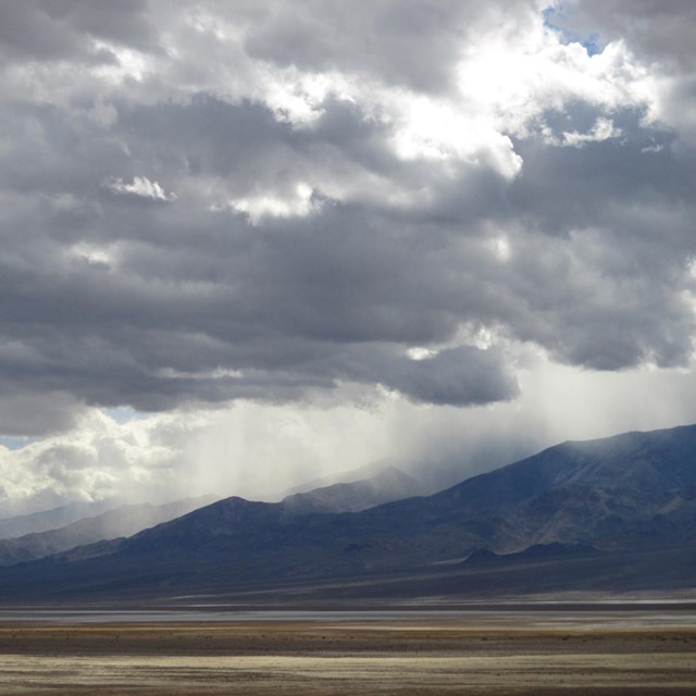Rain falls on mountains from dark clouds in a barren desert landscape.