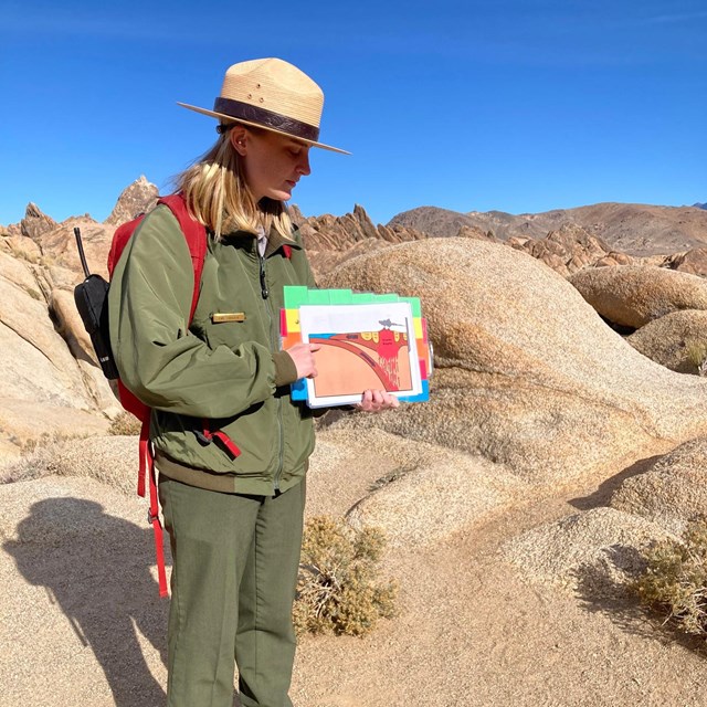 Ranger in uniform stands on large rocks while holding interpretive media.