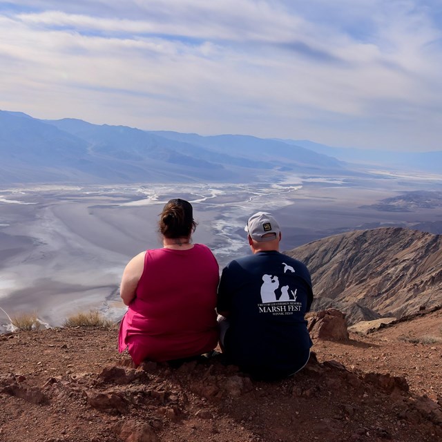 Two individuals sit on the ground, enjoying a view of a salt-covered valley surrounded by mountains.