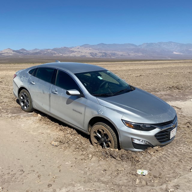 Silver car with two front tires stuck in mud on a valley floor.