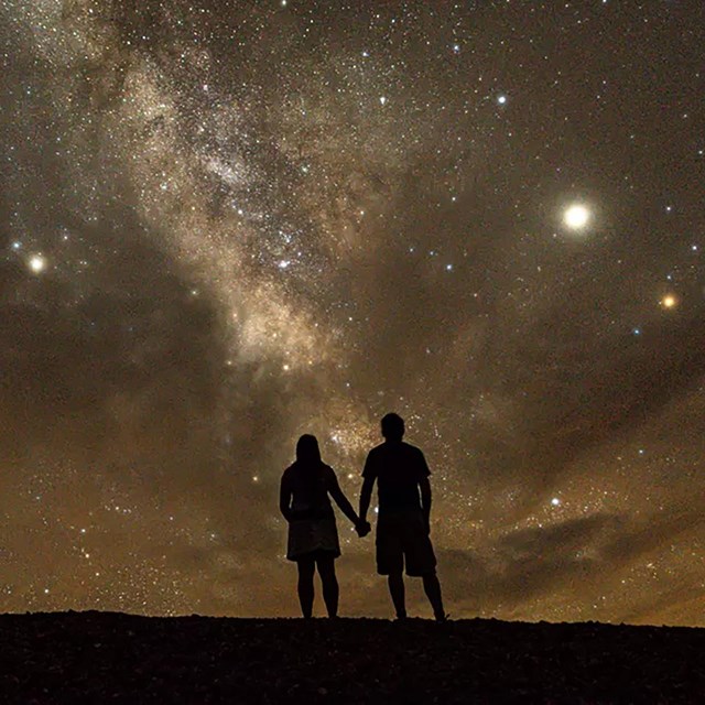 Two silhouetted figures stand holding hands looking into a night sky filled with stars.