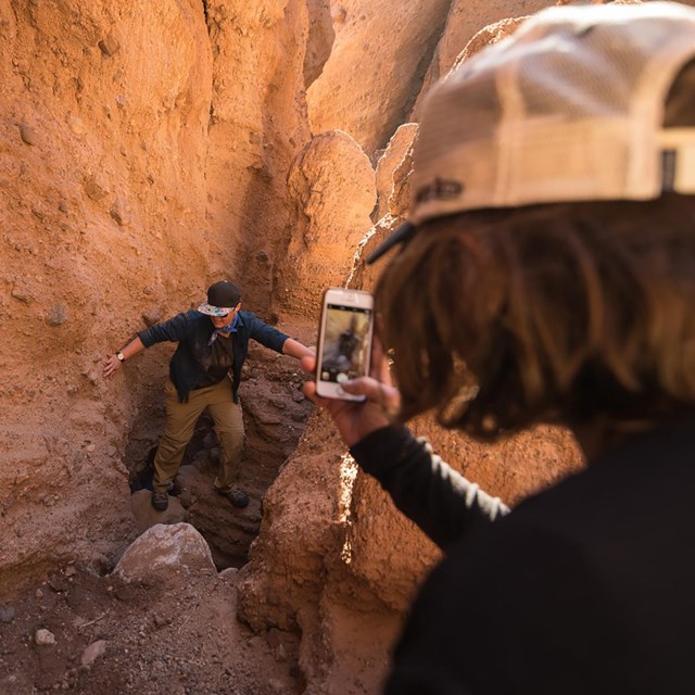 A visitor photographs another visitor hiking in a golden-walled canyon.