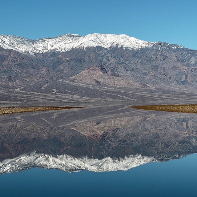 Snowcapped mountains reflected on still water with a clear blue sky.