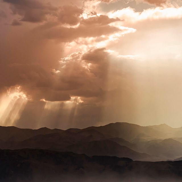 Clouds break and let light through, towering over mountain silhouettes.