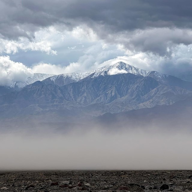 Clouds of sands blowing in the wind along a valley floor with snow capped mountains.