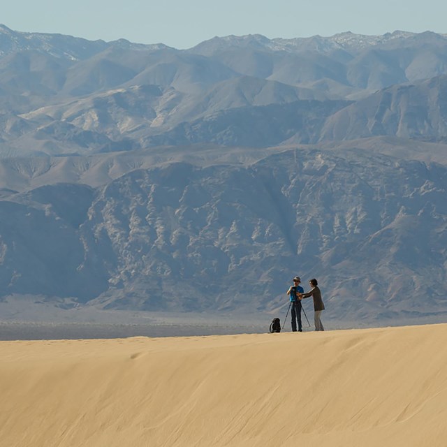Photographers stand atop a sand dune with a brown mountain landscape in the background.