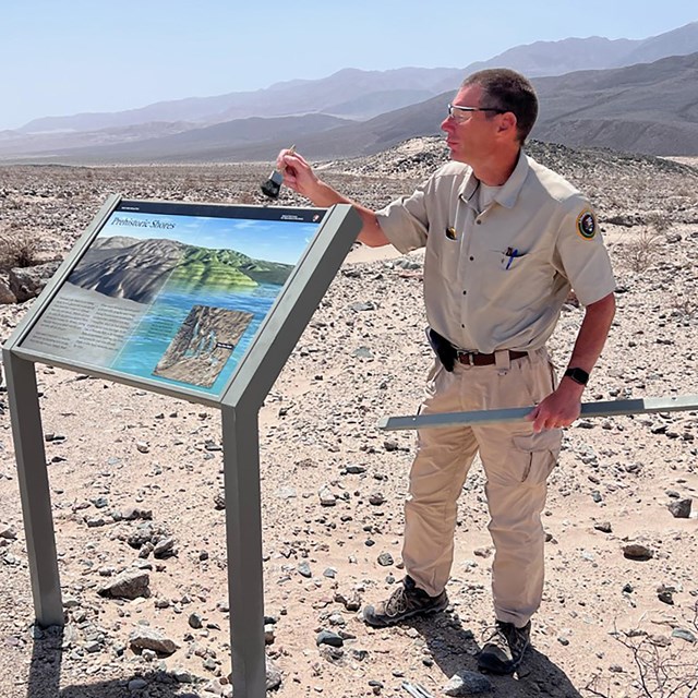 Volunteer Jim in uniform, dusting off a brand-new informational wayside he installed in the desert