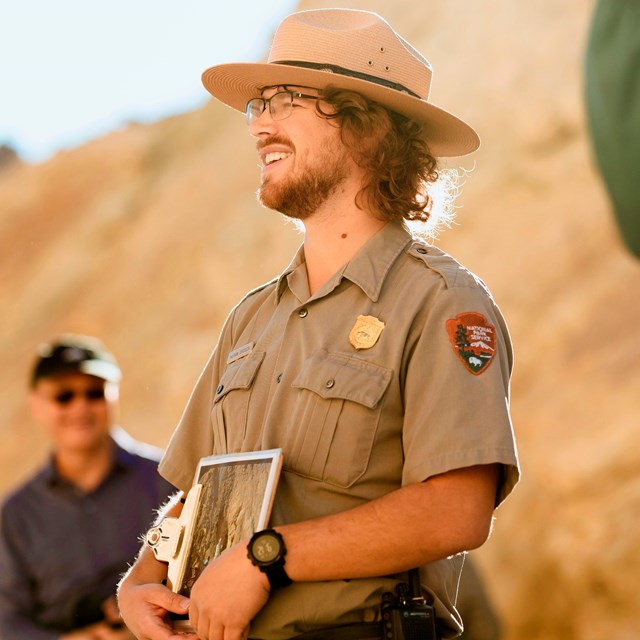 Uniformed ranger holds a clipboard while addressing a group in a yellow canyon.