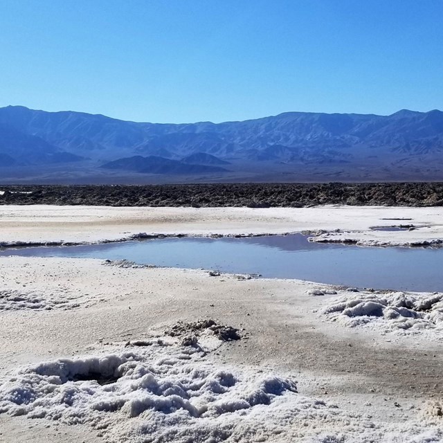 A pool of water is surrounded by a broad, flat valley covered in white, crystalized mineral deposits