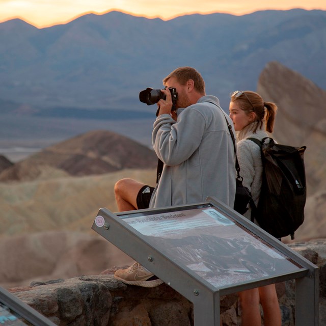 Two visitors looking over hills and mountains while one visitor takes photos.