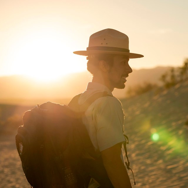 A ranger in uniform wearing a black backpack, smiles with the sun setting behind him over mountains.
