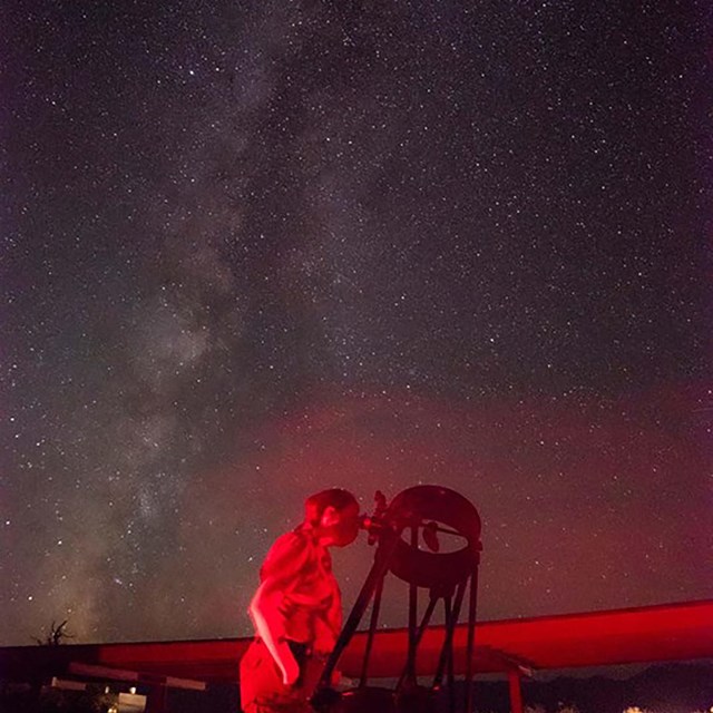 An individual looks into an open-framed telescope with a starry sky behind them.
