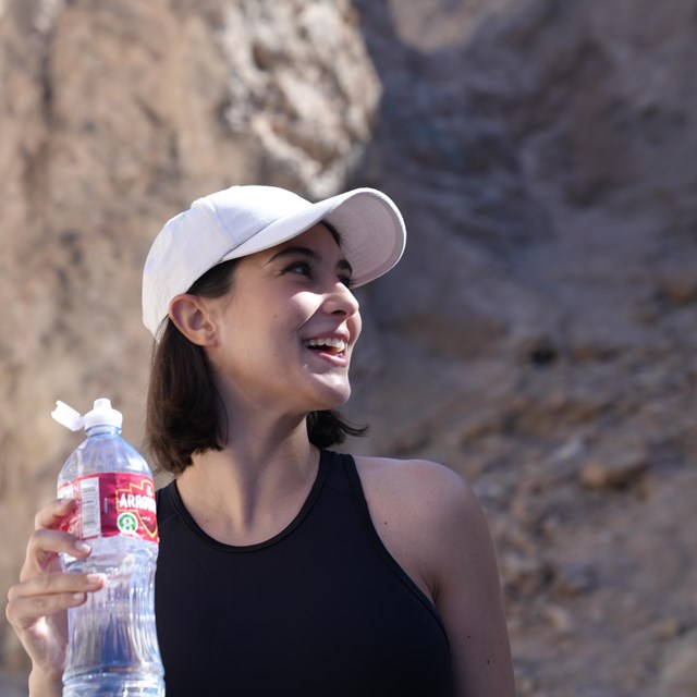 A woman wearing a hat smiles and drinks water while hiking at Golden Canyon.